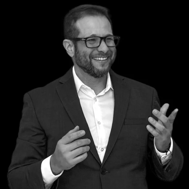 Jay Anaya smiling and gesturing mid-conversation, in a dark suit and glasses